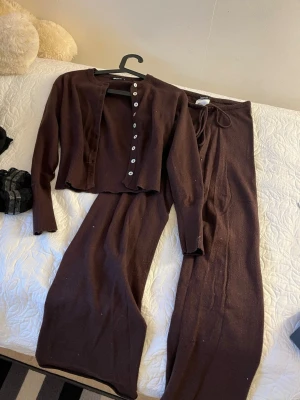 Brunt stickat loungewear-set - Mysigt tvådelat loungewear-set i mörkbrunt stickat material. Setet består av en långärmad kofta med vita knappar och ribbade muddar samt ett par vida byxor med dragsko i midjan. Perfekt för chill dagar. Seten är endast använd en gång men har ett litet fel på armen precis som jag visar på bilden annars är den helt fel fri