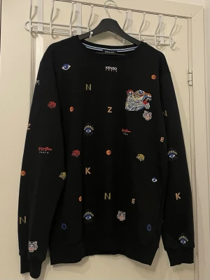 Svart sweatshirt med brodyr från Kenzo - Cool svart sweatshirt från Kenzo med broderade detaljer som ögon, bokstäver och tigrar i flera färger. Rund halsringning och ribbade muddar. Märket syns både på bröstet och i nacken. Perfekt för dig som gillar statement-plagg och streetwear.