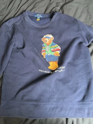 Mörkblå sweatshirt Polo Bear Ralph Lauren - Mörkblå sweatshirt från Ralph Lauren med ikoniska Polo Bear-trycket framtill. Björnen har färgglada kläder och texten 'POLO BEAR BY RALPH LAUREN' under. Tröjan har rund hals och ribbade muddar. Perfekt för dig som gillar klassisk streetwear med en twist.