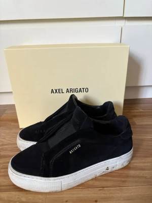 Svarta sneakers Axel Arigato - Stilrena svarta sneakers från Axel Arigato i mocka med elastisk detalj över vristen och vit sula. Diskret logga i guld på sidan. Perfekta för dig som gillar minimalistisk och modern design. Dom är i storlek 37 och tvättas självklart innan fraktning. Väldigt bra skick! 