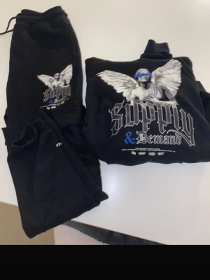 Svarta mjukissett Supply & Demand - Säljer ett svart sett från Supply & Demand med tryck av en ängel och texten 'Supply & Demand' i vitt och blått på ena benet. Byxorna har resår i midjan och mudd vid benslut. Perfekta för en avslappnad och trendig look.