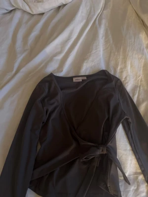 Svart omlottopp med knytband - Säljer en svart omlottopp från NA-KD med långa ärmar och knytband i midjan. Toppen har en djup v-ringning och är tillverkad i ett mjukt, stretchigt material som sitter snyggt på kroppen. Perfekt för dig som gillar stilrena och trendiga plagg.