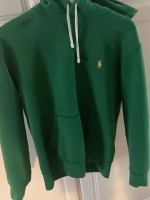 Grön hoodie från Polo Ralph Lauren - Snygg grön hoodie från Polo Ralph Lauren med klassisk gul broderad logga på bröstet. Hoodien har vit dragsko i huvan och en stor magficka framtill. Tillverkad i mjukt material som känns skönt mot huden. Perfekt för en avslappnad och trendig look.