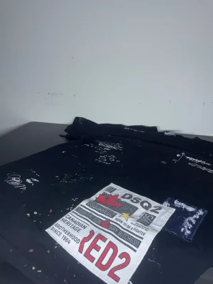 Svart DSQUARED2 t-shirt med patch - Cool svart t-shirt från DSQUARED2 med stor patch framtill, tryckta texter och färgstänk för en edgy look. T-shirten har korta ärmar och rund hals. Perfekt för dig som vill sticka ut med streetwear-vibe.