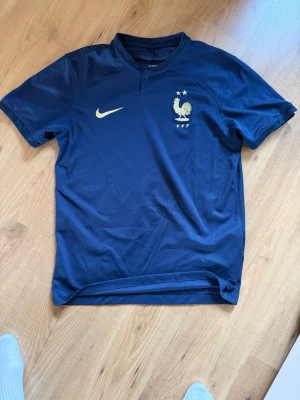 Blå Frankrike fotbollströja från VM 2022 - Mörkblå fotbollströja från Nike med Frankrikes landslagsmärke och två stjärnor på bröstet. Tröjan har korta ärmar, rundad hals med knapp och är tillverkad i ett lätt, andningsbart material perfekt för träning eller match. Pris kan diskuteras vid snabb affär.