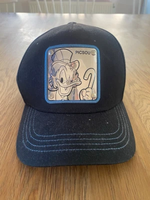 Blå keps med Picsou-motiv från Capslab - Cool blå keps från Capslab med ett stort patch på fronten med Picsou-motiv. Kepsen har vit kontrastsöm på skärmen och justerbar baksida. Materialet är bomull och designen är klassisk med böjd skärm. Perfekt för dig som gillar Disney och unik streetstyle.