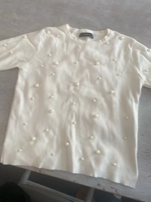 Vit tröja med pärldetaljer - Trendig vit tröja med rund hals och dekorativa pärlor över hela framsidan. Tröjan har en rak passform och långa ärmar, perfekt för att lyfta vilken outfit som helst med en elegant touch.