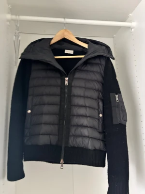 Moncler cardigan Storlek S - Snygg svart pufferjacka med stickade ärmar och vadderad kropp. Jackan har dragkedja framtill, två fickor med tryckknappar och en ficka med dragkedja på ärmen. Utrustad med en stor vadderad huva och ribbade muddar. Perfekt för kalla vinterdagar. Få snittar har gått av vid höger ärmen,  men syns inte. Kvitto saknas 
