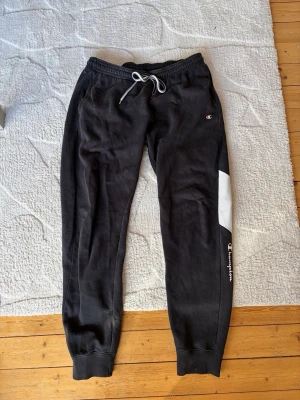 Svarta mjukisbyxor från Champion - Säljer ett par svarta mjukisbyxor från Champion med vit detalj på ena benet och logga tryckt längs benet. Byxorna har resår i midjan med dragsko och muddar vid bensluten. Perfekta för chill eller träning.