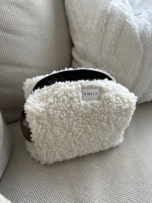 Vit fluffig necessär från Emelo - Supersöt och mjuk necessär från Emelo i vit teddy/fårskinnsimitation. Rektangulär form med brunt handtag på sidan och dragkedja upptill. Perfekt för smink eller småprylar när du är på språng. Storlek medium. Mått ca 20x12x8cm 