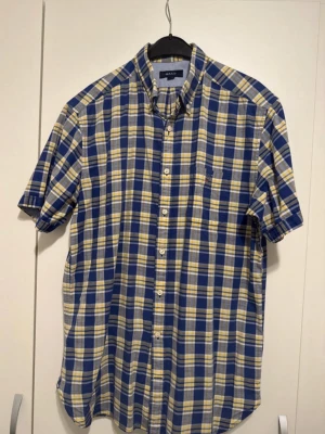 Blårutig kortärmad skjorta Gant - Kortärmad skjorta från Gant i blått, vitt och gult rutmönster. Klassisk krage och knappar framtill. Tillverkad i mjuk bomull som känns skön mot huden. Perfekt för en avslappnad och stilren look.