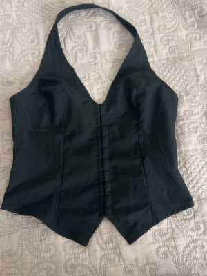 Svart halterneck korsettopp - Snygg svart korsettopp med halterneck och djup v-ringning. Toppen har en spetsig nederkant framtill och markerade sömmar för en figurnära passform. Perfekt att styla med jeans eller kjol för en trendig look.