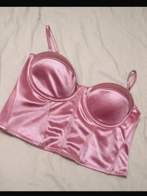 Rosa satin bustier med smala band - Snygg rosa bustier i glansigt satinmaterial med vadderade kupor och smala axelband. Toppen har en figurnära passform och dragkedja bak för enkel påklädning. Perfekt för att skapa en trendig och feminin look.