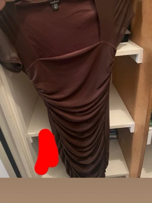 Brun  klänning Boohoo - Säljer en brun midiklänning från Boohoo med snygga draperingar framtill. Klänningen har korta ärmar och en figurnära passform.