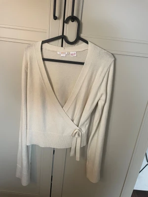 Beige omlottkofta från Gina Tricot - Snygg beige stickad omlottkofta från Gina Tricot med knytband i midjan. Koftan har lång ärm, djup v-ringning och bred ribbad kant nedtill och vid ärmsluten. Perfekt att styla över ett linne eller t-shirt för en trendig look.
