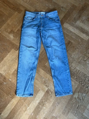 Blå raka jeans i klassisk modell - Säljer ett par blå jeans med rak passform och hög midja storlek i W/L uppskattas till W29/32L. Jeansen har fem fickor, bälteshällor och stängs med dragkedja och knapp. Materialet är klassisk denim i bomull och färgen är ljusblå med lätt tvättad look. Perfekta till en avslappnad stil.