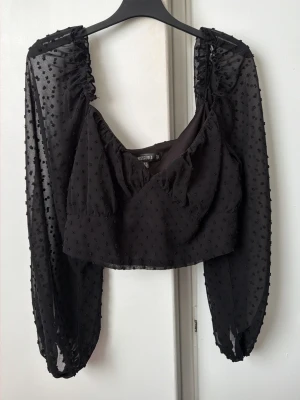 Svart prickig croppad topp Missguided - Säljer en svart croppad topp från Missguided med transparenta långärmar och små prickiga detaljer. Toppen har v-ringning, volang vid ringningen och är tillverkad i ett lätt, syntetiskt material. Perfekt för dig som vill ha en trendig och luftig look. Skulle säga att den är mindre i storleken