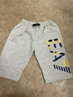 Grå EA7 shorts med tryck - Snygga grå shorts från EA7 med elastisk midja och snörning. Stora blå och gula tryckdetaljer på ena benet ger en sportig vibe. Perfekta för sommardagar eller chill hemma. Tillverkade i mjuk bomull för maximal komfort.