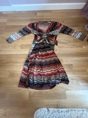 Mönstrad långklänning med knyt - Säljer en långklänning med vågigt mönster i vinrött, svart, beige och vitt. Klänningen har lång ärm, v-ringning och ett snyggt knyt i midjan. Perfekt för dig som gillar färg och retrovibbar!
