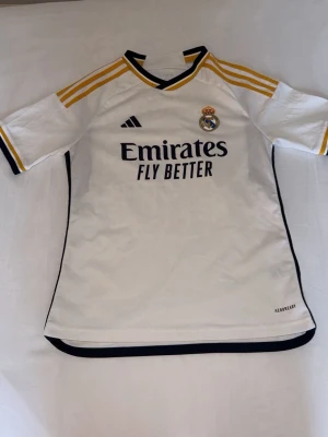 Real Madrid matchtröja Adidas vit - Säljer en vit Real Madrid matchtröja från Adidas med gula och svarta detaljer. Tröjan har klubbmärke och Adidas-logga på bröstet samt 'Emirates Fly Better' tryckt framtill. Den är äkta från Adidas, i storlek 164