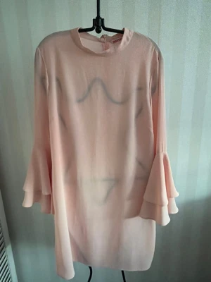 Ljusrosa volangklänning från Indiska - Supersöt ljusrosa klänning från Indiska med volangärmar och rak passform. Klänningen har rund halsringning och är tillverkad i ett lätt, skönt material som ger ett flowy intryck. Perfekt för dig som gillar romantiska detaljer och en feminin look.