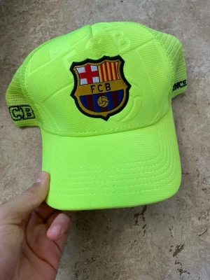 Neongul FC Barcelona keps - Snygg neongul keps med FC Barcelona-logga framtill. Kepsen har mesh på sidorna och justerbar snapback baktill. Broderade detaljer och klubbnamn på insidan. Perfekt för dig som vill sticka ut och visa vilket lag du hejar på.