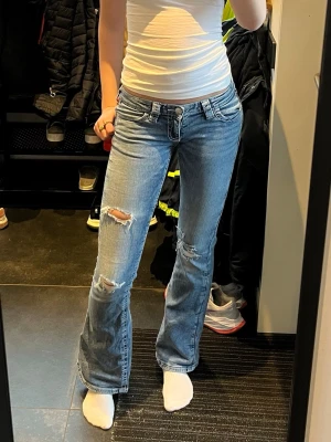 Bootcut jeans med slitningar - Säljer ett par ljusblå bootcut jeans med slitna detaljer och hål på benen. Jeansen har låg midja, klassiska fem fickor och dragkedja med knapp. Perfekta för en avslappnad och trendig look.