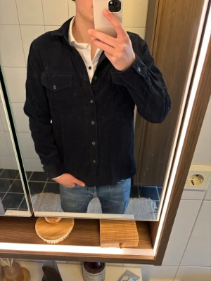 Mörkblå overshirt från Dobber - Säljer en stilren mörkblå overshirt från Dobber i mjuk manchester. Skjortan har två bröstfickor med knappar, klassisk krage och knäppning framtill. Perfekt att bära över en t-shirt eller skjorta för en avslappnad men snygg look.