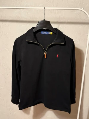 Svart stickad tröja Polo Ralph Lauren - Svart stickad tröja från Polo Ralph Lauren med halv dragkedja och klassisk röd logga broderad på bröstet. Tröjan har hög krage och långa ärmar, perfekt för lager på lager. Materialet känns mjukt och bekvämt.