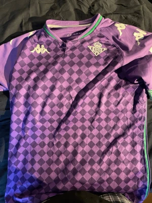 Lila Real Betis Fotbollströja Kappa L - Säljer en lila och mörklila rutmönstrad matchtröja från Real Betis, tillverkad av Kappa. Tröjan har vita Kappa-loggor på axlarna, klubbmärke på bröstet och gröna detaljer vid kragen. Perfekt för dig som älskar fotboll och vill sticka ut på planen.