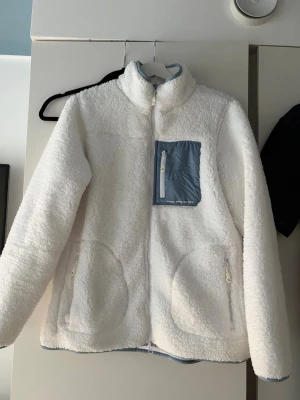 Vit teddyjacka med blå detalj - Mysig vit teddyjacka med hög krage och dragkedja framtill. Jackan har en blå bröstficka med vit dragkedja och två sidofickor med dragkedja. I stl 40/42 se bild.