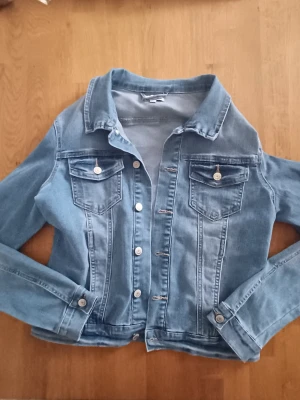 Ljusblå klassisk jeansjacka - Snygg ljusblå jeansjacka med raka sömmar och två bröstfickor med lock och knapp. Jackan har silverfärgade knappar framtill och vid ärmsluten samt en klassisk krage. Perfekt att slänga över en hoodie eller t-shirt för en avslappnad look.