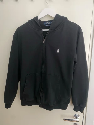Svart polo zip up - Klassisk svart hoodie med dragkedja från Polo Ralph Lauren. Har huva, två stora fickor framtill och den ikoniska vita loggan broderad på bröstet. Tillverkad i  bomullsmaterial och har ribbade muddar vid ärmar och nederkant. Sälja pga att den inte passar mig längre.