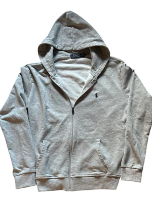 Grå hoodie med dragkedja Polo Ralph Lauren - Ljusgrå hoodie från Polo Ralph Lauren med dragkedja, huva och två stora fickor framtill. Klassisk broderad logga på bröstet och ribbade muddar. Tillverkad i mjukt bomullstyg som känns riktigt skönt.