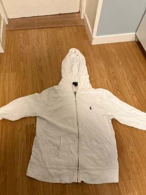Vit hoodie från Polo Ralph Lauren - Säljer en vit hoodie från Polo Ralph Lauren i storlek L (14-16). Är 170 och sitter som S. Hoodien har dragkedja, två fickor framtill och den klassiska Polo-loggan broderad på bröstet. Tillverkad i mjukt bomullsmaterial och har en skön huva. Perfekt för en chill och stilren look.
