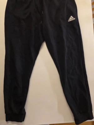 Svarta Adidas mjukisbyxor - Svarta mjukisbyxor från Adidas med vit broderad logga. Tillverkade i mjukt material, troligtvis bomullsblandning. Perfekta för chill eller träning och hur sköna som helst       Midja: 40 cm  och stretchig        Benlängd  68 cm      Hela   98 cm    passar L också