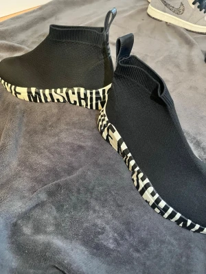 Moschino skor - Svarta sock sneakers från Moschino med stretchig stickad ovandel och vit sula med svart textprint runtom. Skorna har en strömlinjeformad siluett och pull tab bak för enkel påtagning. Perfekta för en trendig streetwear-look.