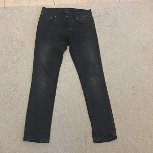 Svarta jeans från Nudie Jeans - Snygga grim tim jeans svart/gråa från Nudie Jeans med klassisk femficksdesign och subtila slitningar. Jeansen har rak passform och är tillverkade i mjukt denimtyg med svarta sömmar och Nudie Jeans-logga på bakfickan. Perfekt för dig som gillar stilrena och tidlösa jeans. Använt fåtal gånger och har kvar klädlapparna.