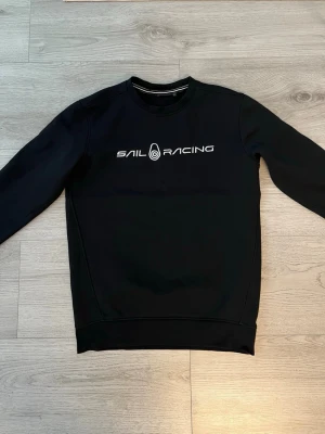 Svart Sail Racing sweatshirt - Stilren svart sweatshirt från Sail Racing med vitt tryck på bröstet. Tröjan har rund halsringning, långa ärmar och ribbade muddar vid ärmslut och nederkant. Perfekt för en avslappnad och sportig look.