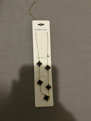 Guldhalsband med svarta fyrklöver - Snyggt halsband i rostfritt stål med guldfärgad kedja och fem svarta fyrklöverformade hängen. Kan justeras i längd och har små pärldetaljer runt varje hänge. Perfekt accessoar för att lyfta din outfit. Aldrig använt 