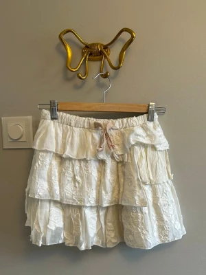 Vit volangkjol - Söt vit kortkjol med flera volanger. Kjolen har broderade detaljer och är perfekt för sommarens alla tillfällen. Tillverkad i ett lätt och luftigt material.