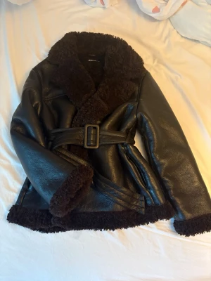 Teddy PU belted jacket - Säljer en teddyfodrad belted PU skinnjacka från Gina Tricot som inte säljs längre.  Snygg PU-finish (konstläder) i mörkbrunt/svart med ett varmt och mysigt teddyfoder i chokladbrunt. Skärp i midjan för en fin silhuett.  Storlek XS, men passar även S. Aldrig använd, har endast hängt i förrådet.  Pris kan diskuteras.