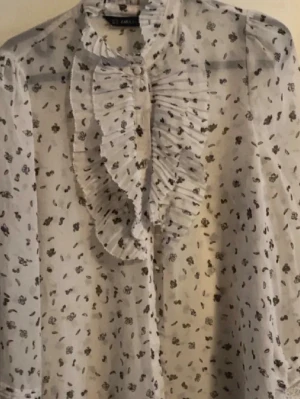 Vit blus med volang och småblommigt - Söt vit blus från Zara med små blommönster i svart och volangdetaljer längs knäppningen framtill. Blusen har lång ärm och är gjord i ett tunt, transparent material som ger en luftig känsla. Perfekt för en trendig och romantisk look.