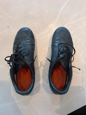 Svarta Nike phantom fotbollsskor - Säljer ett par svarta Nike fotbollsskor med snörning och orange innersula. Skorna har en modern, strömlinjeformad design och slitstark ovandel i syntetmaterial. Dobbarna är svarta med orange detaljer, perfekta för spel på gräsplan.