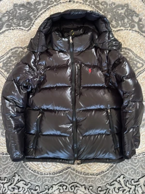 Svart dunjacka från Polo Ralph Lauren - Snygg svart dunjacka från Polo Ralph Lauren med glansig finish och röd broderad logga på bröstet. Jackan har huva, dragkedja framtill och flera praktiska fickor med dragkedja. Perfekt för kalla vinterdagar och riktigt stilren look.