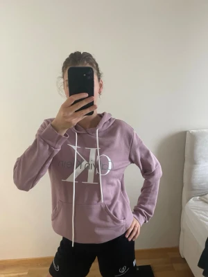 Ljuslila hoodie från Calvin Klein - Säljer en ljuslila hoodie från Calvin Klein med stor vit CK-logga framtill. Hoodien har huva med vita dragsnören och en klassisk känguruficka. Mjuk och skön bomullsblandning, perfekt för chill dagar. Snygg och enkel design som passar till det mesta.