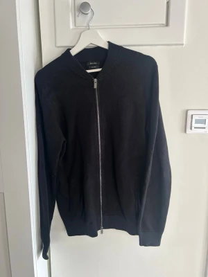 Svart ziptröja från Massimo Dutti - Stilren svart ziptröja från Massimo Dutti med hel dragkedja framtill och ribbade muddar vid ärmslut och nederkant. Tröjan har en klassisk passform och rund halsringning, perfekt att slänga på sig över en t-shirt. Materialet känns mjukt och bekvämt.