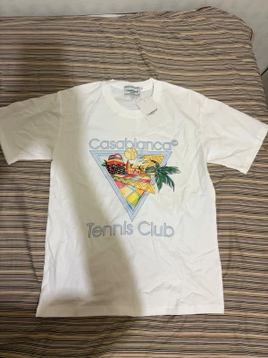 Vit Casablanca Tennis Club t-shirt - Snygg vit t-shirt från Casablanca med färgglatt tryck framtill och texten 'Casablanca Tennis Club'. Klassisk passform och korta ärmar. Perfekt för dig som gillar sportig och trendig stil.