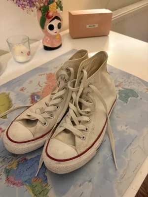 Vita Converse All Star high tops - Klassiska vita Converse All Star high tops med röd rand längs sulan och vit gummitå. Skorna har canvasmaterial, vita skosnören och den ikoniska loggan på sidan. Perfekta för en avslappnad och cool stil.