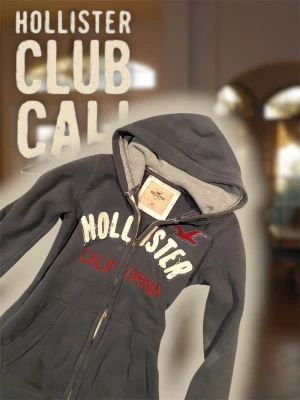 Mörkgrå Hollister hoodie med dragkedja - Snygg mörkgrå hoodie från Hollister med dragkedja (inte orginal så var försiktig) framtill och stor huva. Framsidan har vit och röd brodyr med texten 'Hollister California' samt Hollisters logga i rött. Mjuk insida och två fickor framtill. Perfekt för en avslappnad och cool stil. Det står att den är storlek medium men den sitter som small.
